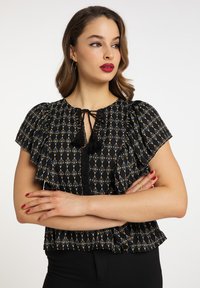 Černá blůza s vzorem, která má krátké volánkové rukávy, detaily na krku na zavázání a třásně. Látka má květinový a geometrický design.