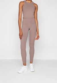 Femme portant un legging taille haute mauve et un crop top sans manches assorti, avec des chaussures de sport blanches, debout devant un fond uni.