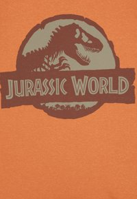 Oransje stoff med et grafisk design som har en dinosaur-siluett og teksten "Jurassic World" i brune og grå farger, i sirkulær form.