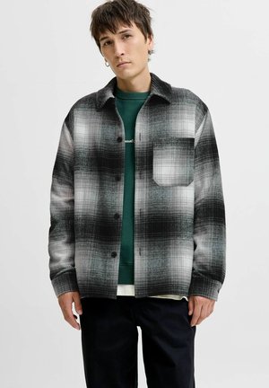 Zwart-wit geruite overhemdjack met knoopsluiting, één borstzak en een losse pasvorm, gedragen over een groene sweatshirt.