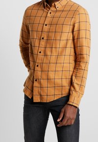Camicia in flanella gialla senape con motivo a quadri neri, colletto button-down, maniche lunghe e cinque bottoni neri sulla parte anteriore.