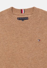 Καφέ πλεκτό πουλόβερ με στρογγυλή λαιμόκοψη και ριμπ κολλέ. Έχει μικρό έμβλημα σε κόκκινο, άσπρο και ναυτικό μπλε χρώμα στο στήθος.