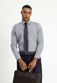 Camisa de rayas azul claro para hombre con cuello afilado, combinada con una corbata oscura y pantalones azul marino; sosteniendo un maletín de cuero marrón.
