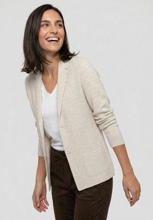 Woman with dark hair wearing a beige cardigan, white shirt, and dark pants standing and smiling while looking slightly to the side.