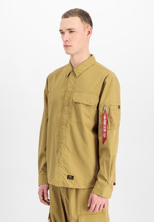 Junger Mann, der ein beige langärmliges Hemd mit Brust- und Ärmeltaschen sowie passende Cargohosen vor weißem Hintergrund trägt.