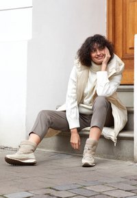 Gilet puffer beige sopra un dolcevita bianco a coste, pantaloni grigi e stivali impermeabili beige chiaro con lacci, seduto sui gradini.