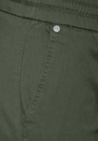 Gros plan sur la poche d'un pantalon vert olive avec des coutures visibles et un détail en rivet métallique près de la taille.