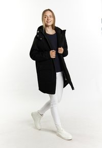 Parka nera con cappuccio, maniche elastiche, zip frontale e grandi tasche, indossata sopra un top scuro e pantaloni bianchi, completata da sneakers bianche.