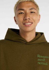 Felpa con cappuccio verde oliva realizzata in tessuto morbido, con cappuccio e testo ricamato in verde: "I corridori non sono normali." Texture liscia.