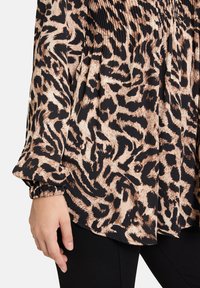 Blouse noire et beige avec un motif léopard, présentant un tissu texturé, des manches longues avec des poignets élastiques et une coupe ample.