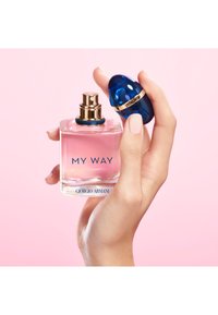 Main tenant un flacon de parfum "MY WAY" de Giorgio Armani rose avec le bouchon bleu retiré sur un fond rose clair.