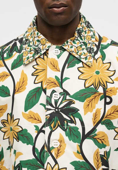 Άνδρας που φοράει λευκό πουκάμισο με μεγάλες κίτρινες, πράσινες και μαύρες λουλουδένιες εκτυπώσεις και κοντράστ γιακά με λουλούδια.