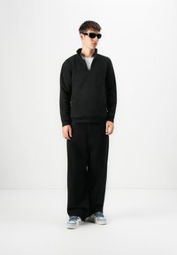 Hombre vestido con un pulóver de forro polar negro, pantalones anchos negros, zapatillas azul y blancas, y gafas de sol negras, contra un fondo blanco liso.