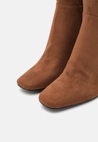 Bottes courtes en daim marron avec un bout pointu, présentant une texture lisse et des coutures minimales pour un aspect épuré et profilé.