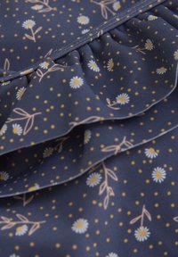 Marinblått tyg med vita prästkragar och gula prickar, med en volangkant och delikata blommönster. Slät textur med subtil lyster.