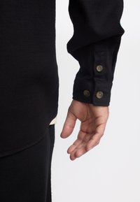 Camisa negra de manga larga, confeccionada con un tejido texturizado, que presenta un puño ajustado con dos botones redondos marrones y un dobladillo crema en capas por debajo.