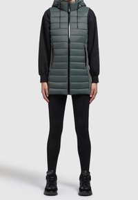 Gilet imbottito verde con zip frontale, cappuccio e due tasche laterali, indossato sopra una maglia nera a maniche lunghe e leggings neri, abbinato a scarpe da ginnastica nere.