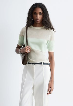 Frau mit lockigem Haar, die ein beige und hellgrünes Kurzarm-Top, weiße hoch taillierte Hose trägt, eine braune Umhängetasche und silberne Ringe.
