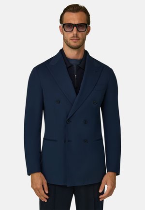 Mand iført sorte solbriller og en dobbeltradet navy blazer over en mørk lynlåstrøje, stående mod en ensfarvet baggrund.