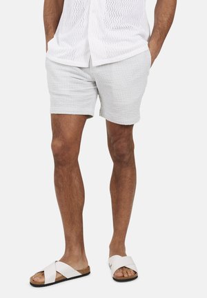 Homme portant un short gris clair texturé, une chemise à manches courtes blanche à motifs, et des sandales blanches à lanières croisées, debout les mains dans les poches.