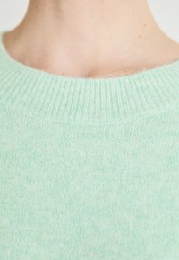 Närbild av en persons hals och övre bröst som bär en mjuk, ljusgrön stickad tröja med ribbat halsringning.