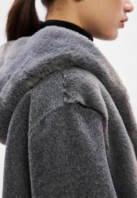 Sivý kabát s kapucňou z umelého kožušiny, s textúrovaným povrchom a hladkým kontrastným golierom. Tkanina je na pohľad mäkká a teplá.
