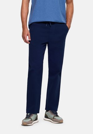 Hombre con pantalones azul marino de corte recto, camiseta azul jaspeada y zapatillas gris-verde, de pie con las manos en los bolsillos.