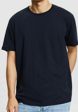 Uomo che indossa una maglietta nera a maniche corte con scollo tondo e jeans azzurri, mostrato dal collo ai fianchi su sfondo semplice.