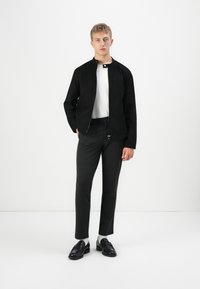 Jeune homme debout portant une veste noire zippée, une chemise blanche, un pantalon foncé, des chaussettes blanches et des mocassins noirs devant un fond blanc uni.