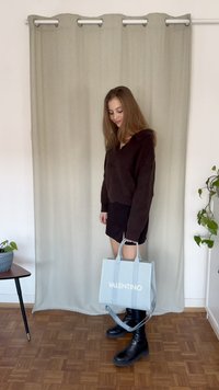 Brūns korduroja džemperis, melna mini svārki; tur rokās gaiši zils VALENTINO somu. Melni augstpapēžu zābaki līdz ceļgaliem. Fons: bēša aizkars un augi.