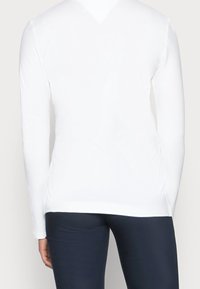 Hvid langærmet skjorte med en glat tekstur, tætsiddende design og en rund kant, båret over mørkfargede leggings.
