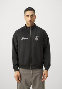 Sort lynlåsjakke med ståkrave, der har et hvidt "Benfica" logo og emblem på brystet. Glat stof, ribbede manchetter.