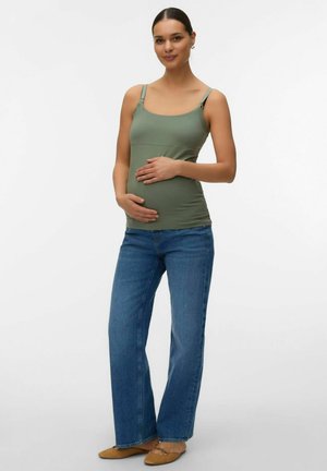 Zwangere vrouw die een groen hemd draagt, blauwe spijkerbroek en bruine platte schoenen, staand met handen zacht op de buik tegen een witte achtergrond.