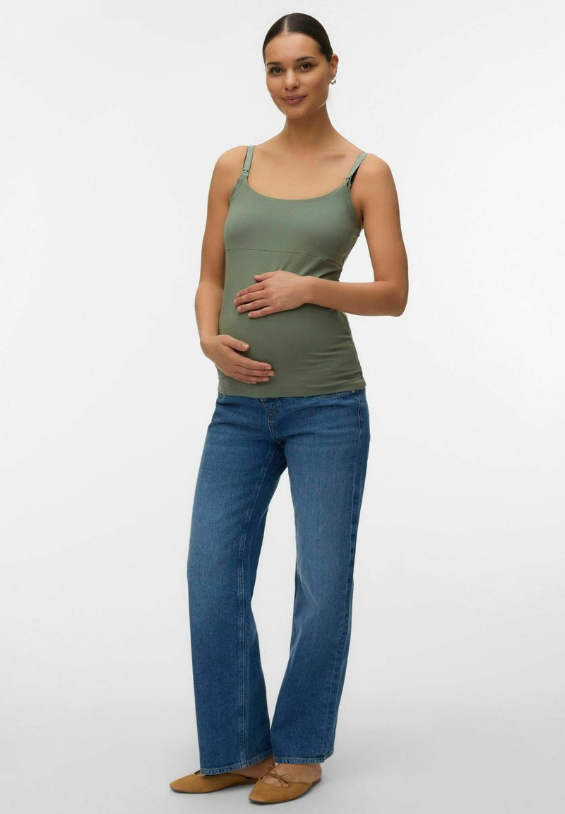 Femme enceinte portant un débardeur vert, un jean bleu et des ballerines marron, debout avec les mains posées délicatement sur le ventre, sur fond blanc.