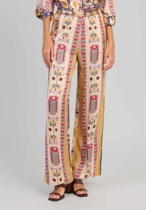 Femme portant un pantalon à jambes larges avec des motifs floraux verticaux en rose, jaune et marron, associé à des sandales marron à talons.