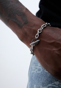 Brăţară din metal argintiu cu închizătoare circulară, având legături intercalate. Purta pe un încheietură cu textură de piele şi tonuri vizibile.