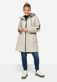 Parka impermeabile grigio chiaro con cappuccio, dettagli neri, due tasche frontali e chiusura con zip, abbinata a jeans blu scuro e stivaletti bianchi.