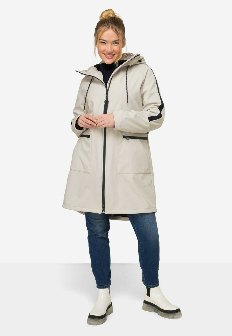 Parka impermeabile grigio chiaro con cappuccio, dettagli neri, due tasche frontali e chiusura con zip, abbinata a jeans blu scuro e stivaletti bianchi.