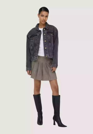 Donna con giacca di jeans nera, top bianco, gonna plissettata grigia e stivali neri alti con tacco, in piedi davanti a uno sfondo neutro.