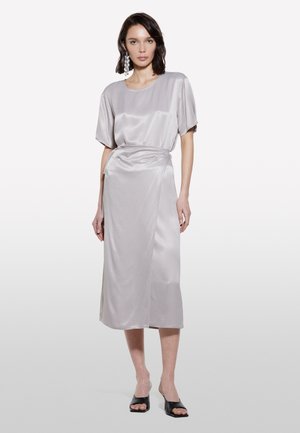 Abito midi in morbido grigio chiaro con vestibilità rilassata, maniche corte e dettaglio con nodo sul lato. Tacchi aperti neri completano l'outfit.