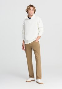 Felpa bianca con zip, pantaloni beige e sneaker bianche con dettagli marroni. Il modello sta in modo casual con una mano in tasca su uno sfondo neutro.