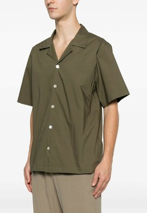 Uomo che indossa una camicia verde oliva a maniche corte con collo ampio e pantaloni beige su sfondo semplice.