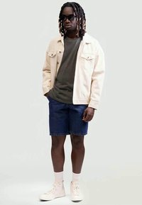 Homem com dreads a usar óculos de sol pretos, casaco bege, camisa verde-escura, calções de ganga, meias brancas e sapatilhas brancas, em pé contra um fundo branco.