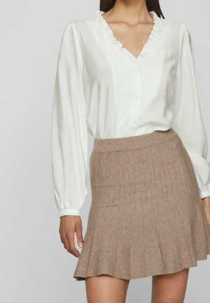 Vrouw draagt een witte blouse met ruches en V-hals, lange mouwen en een hoge taille bruine geplooide rok.