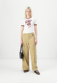 White t-shirt with burgundy trim and "Cherry Coke" graphic, paired with beige cargo pants featuring side pockets, and leopard-print flat shoes.