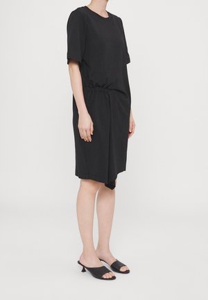 Vrouw draagt een losse zwarte jurk tot op de knie met korte mouwen en zwarte muiltjes met open teen en hak, staand tegen een effen lichte achtergrond.