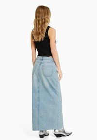 Femme aux longs cheveux ondulés portant un haut sans manches noir, une longue jupe en denim bleu clair et des bottines en argent, se tenant de dos.