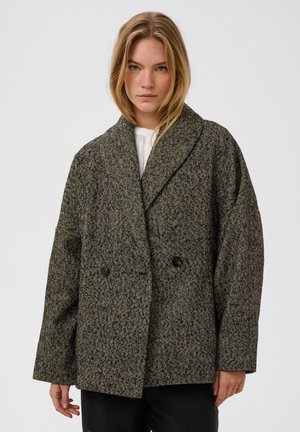 Femme blonde portant un blazer en laine sombre à motif chevrons oversize sur un haut blanc et un pantalon noir, debout devant un fond uni.