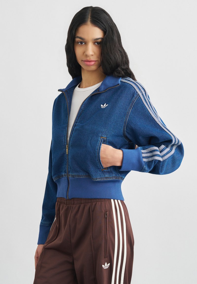 Jachetă din denim cu fermoar, prevăzută cu un guler albastru, două buzunare laterale și accente albe cu trei dungi Adidas pe mâneci. Asortată cu pantaloni maro.