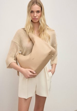 Žena s dlouhými blond vlasy nosí béžový pletený svetr a krémové šortky, drží velkou béžovou koženou tašku přes rameno se zlatými detaily.